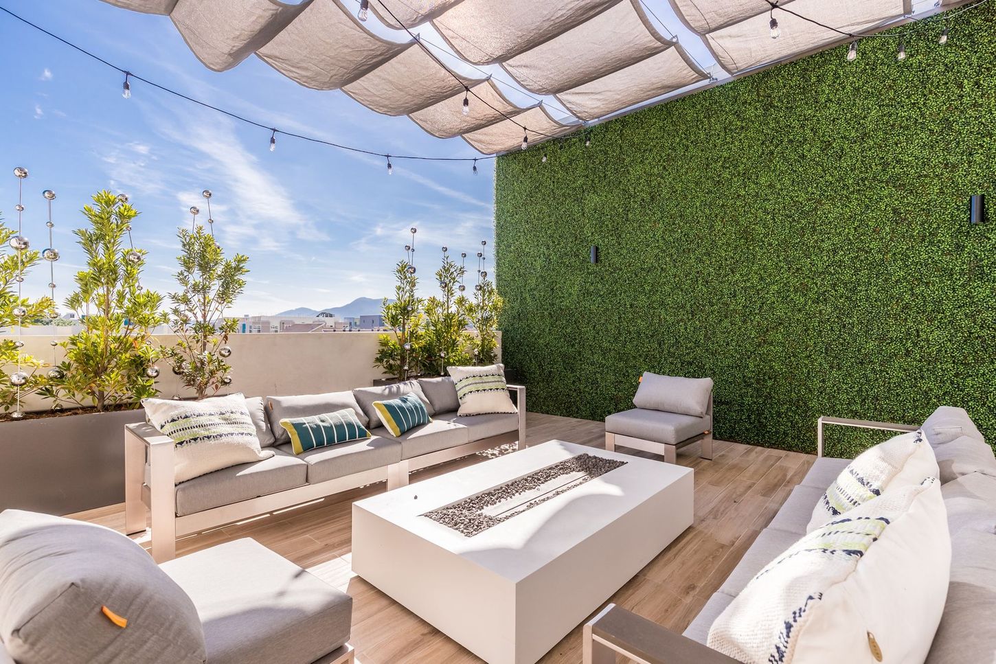 Rooftop patio with white furniture, a fire pit, and a green hedge wall under a fabric canopy.