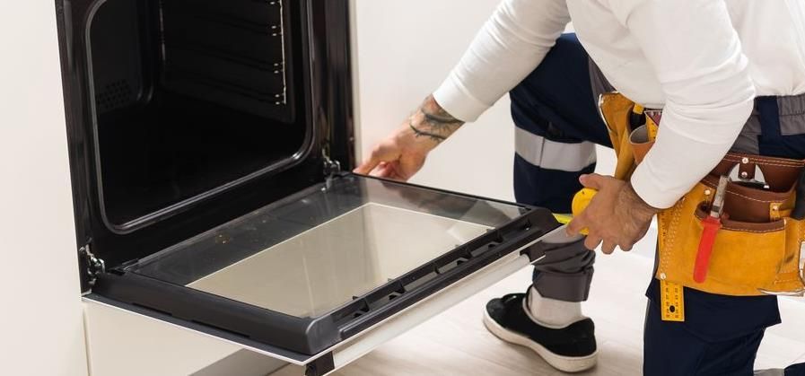 A technician is repairing an oven with the door open, wearing a tool belt.