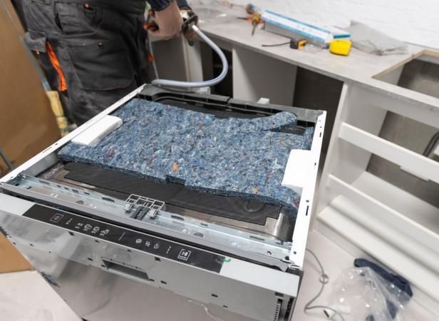 A person installing a dishwasher in a kitchen. The dishwasher is open, showing insulation, with a person using a caulk gun.