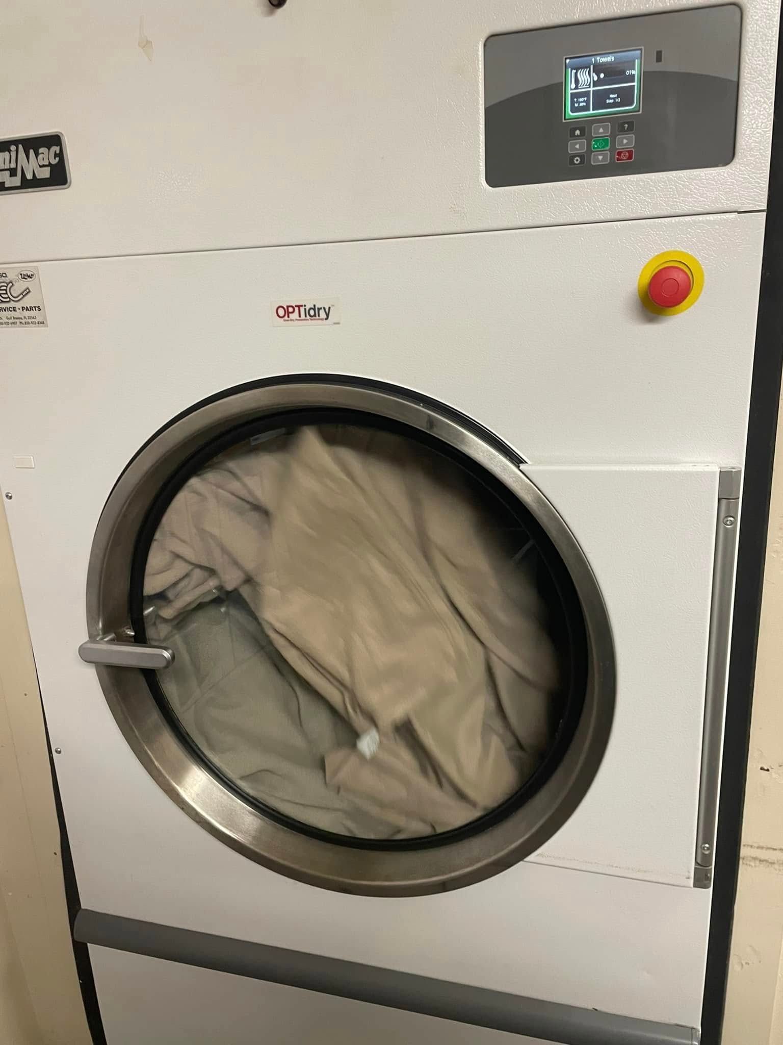 White industrial clothes dryer with clothes inside. Control panel, red stop button.