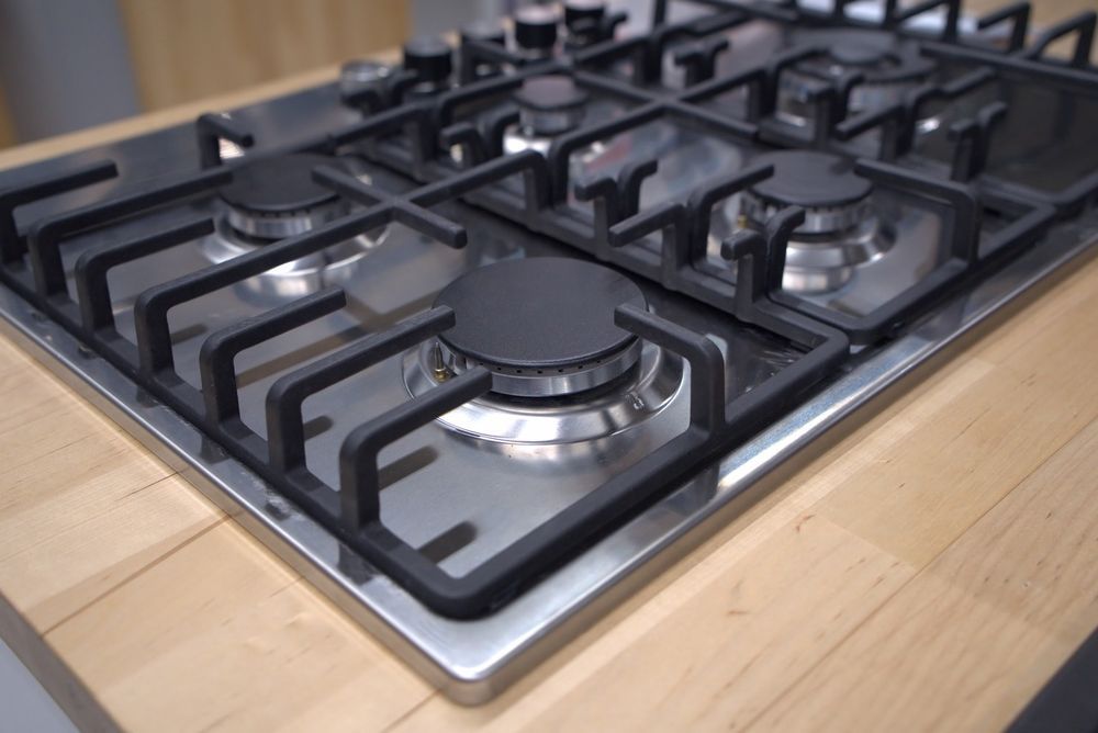 Gas cooktop, stainless steel with black grates and burners, set on a light wood countertop.