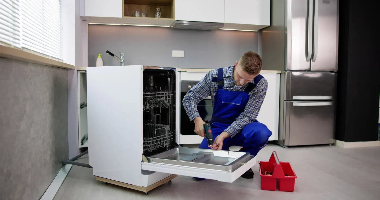 Technician in blue coveralls fixing a dishwasher in a kitchen.