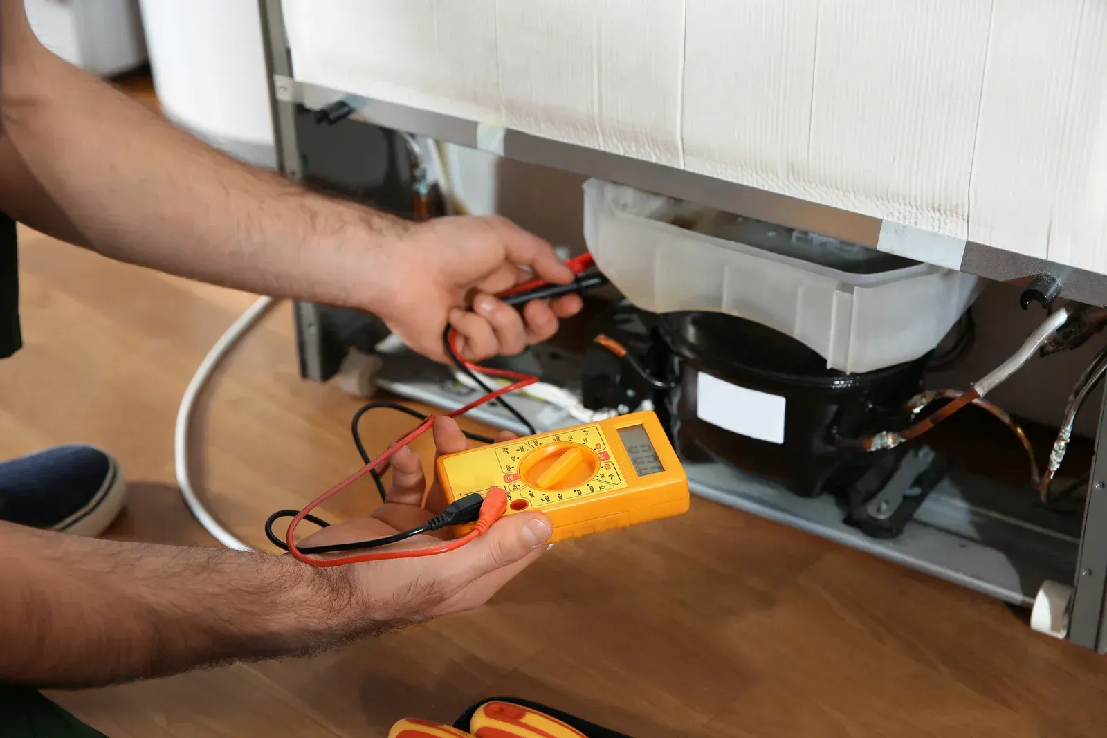 Person using a multimeter to test a refrigerator's compressor; inside a home.