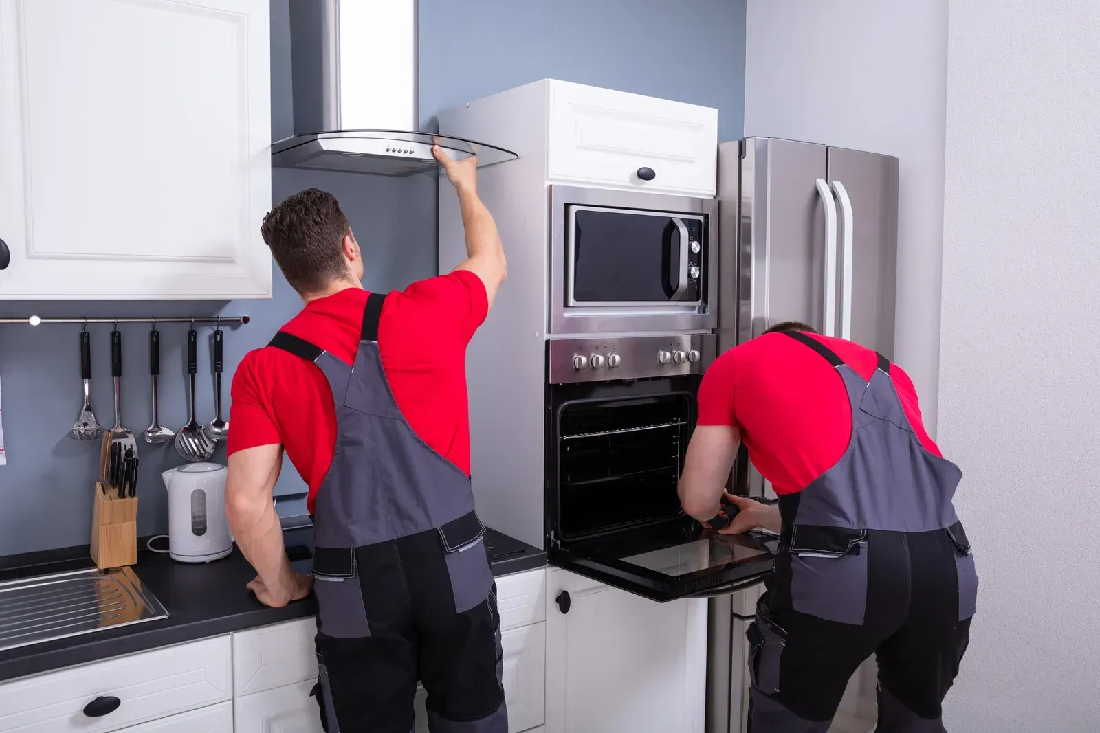 Two workers in red shirts and overalls installing kitchen appliances.
