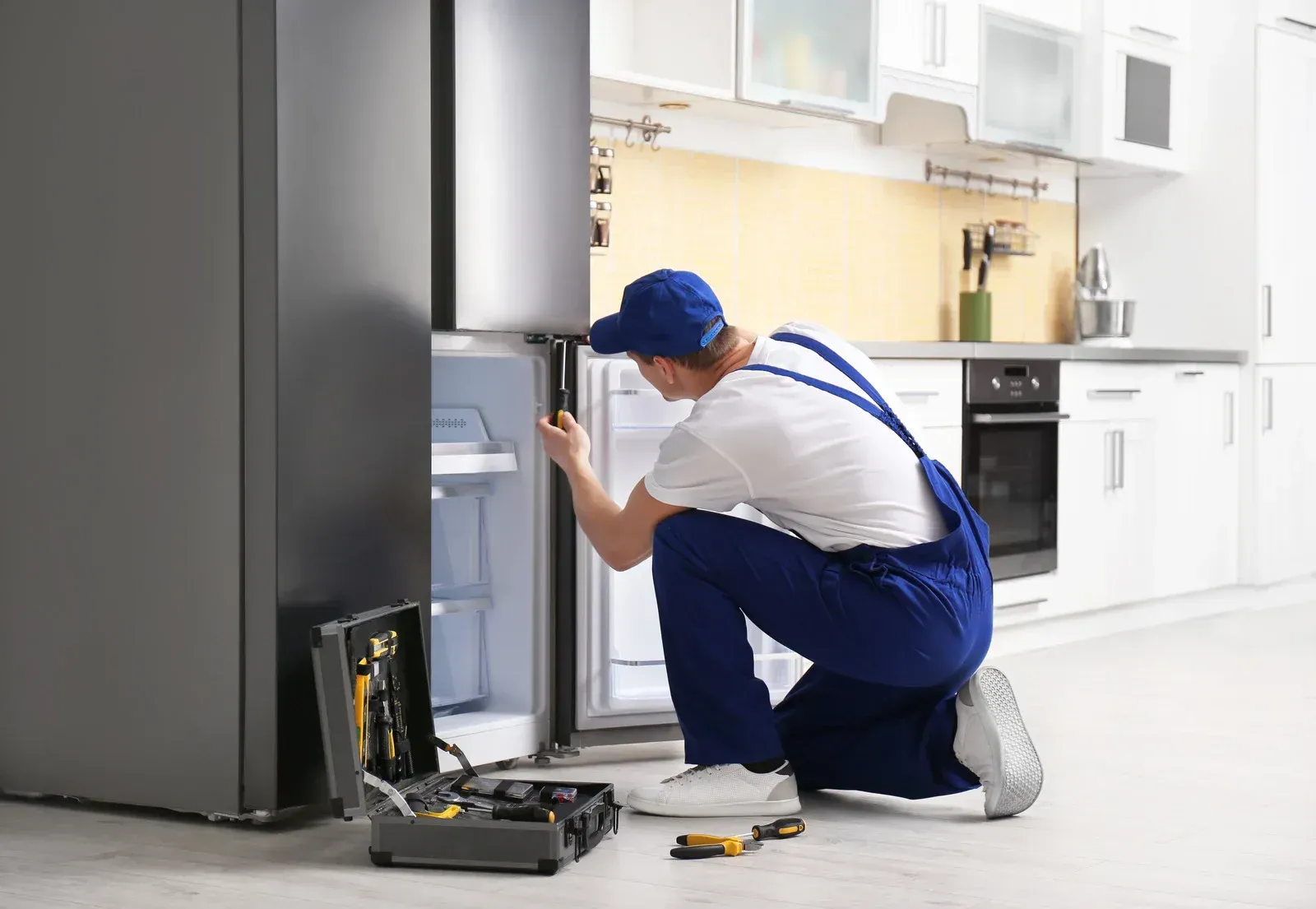 A repair person in blue overalls kneels, fixing a refrigerator in a kitchen with a toolbox open.