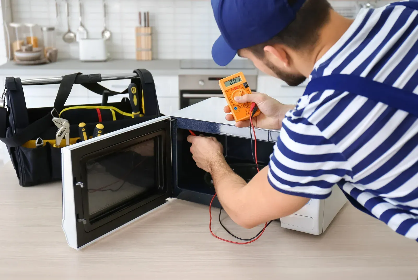Technician testing a microwave with a multimeter; tool bag nearby on a kitchen counter.