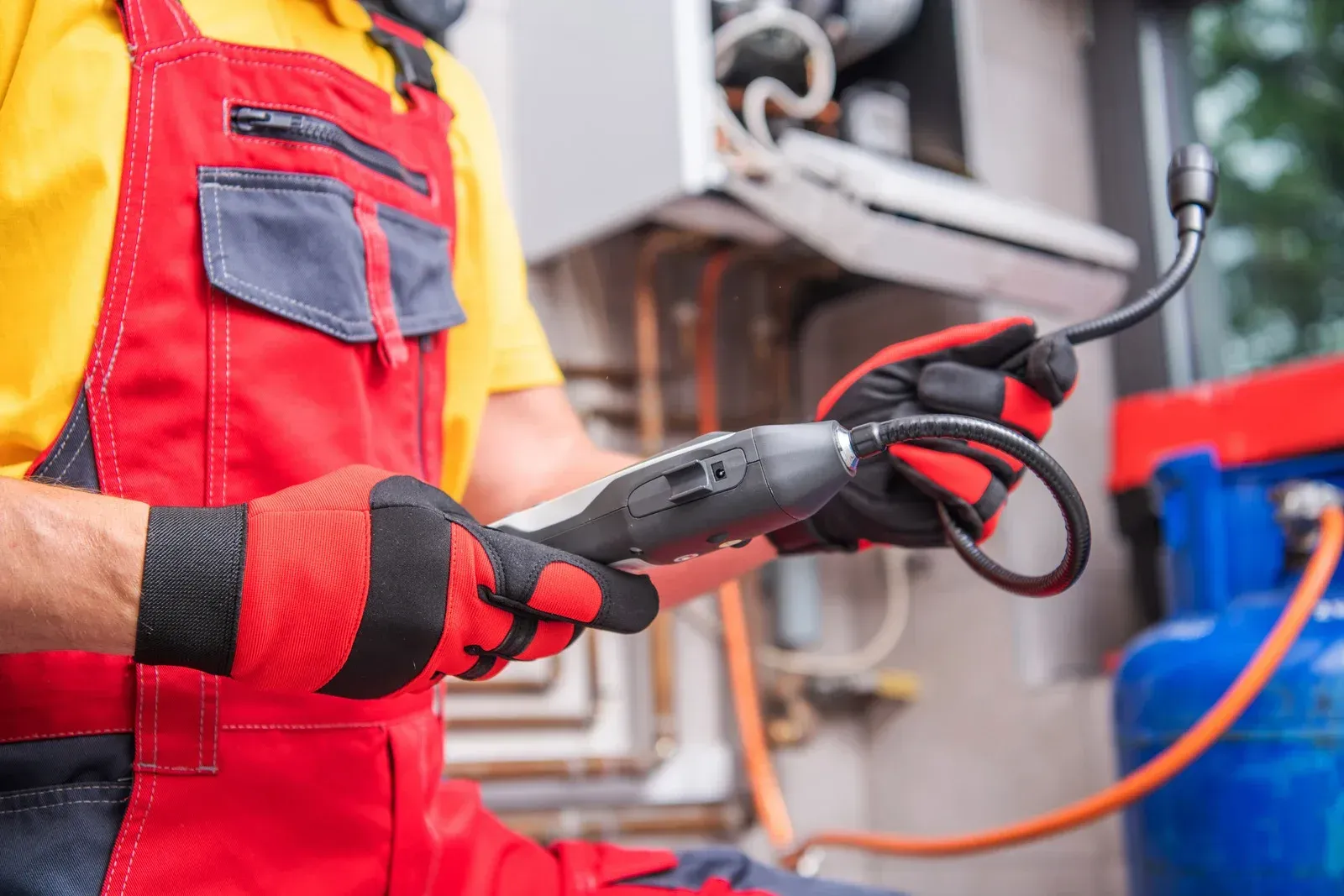 Technician in red overalls and gloves inspecting a gas line near a blue tank and furnace.
