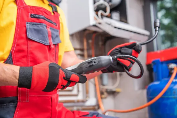 Technician in red overalls and gloves inspecting a gas line near a blue tank and furnace.