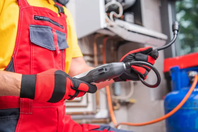 Technician in red overalls and gloves inspecting a gas line near a blue tank and furnace.