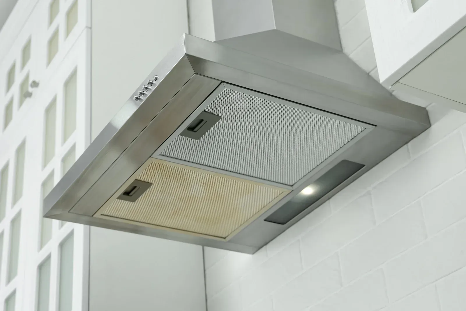 Stainless steel range hood with two filters and a light, mounted above a white tiled wall.