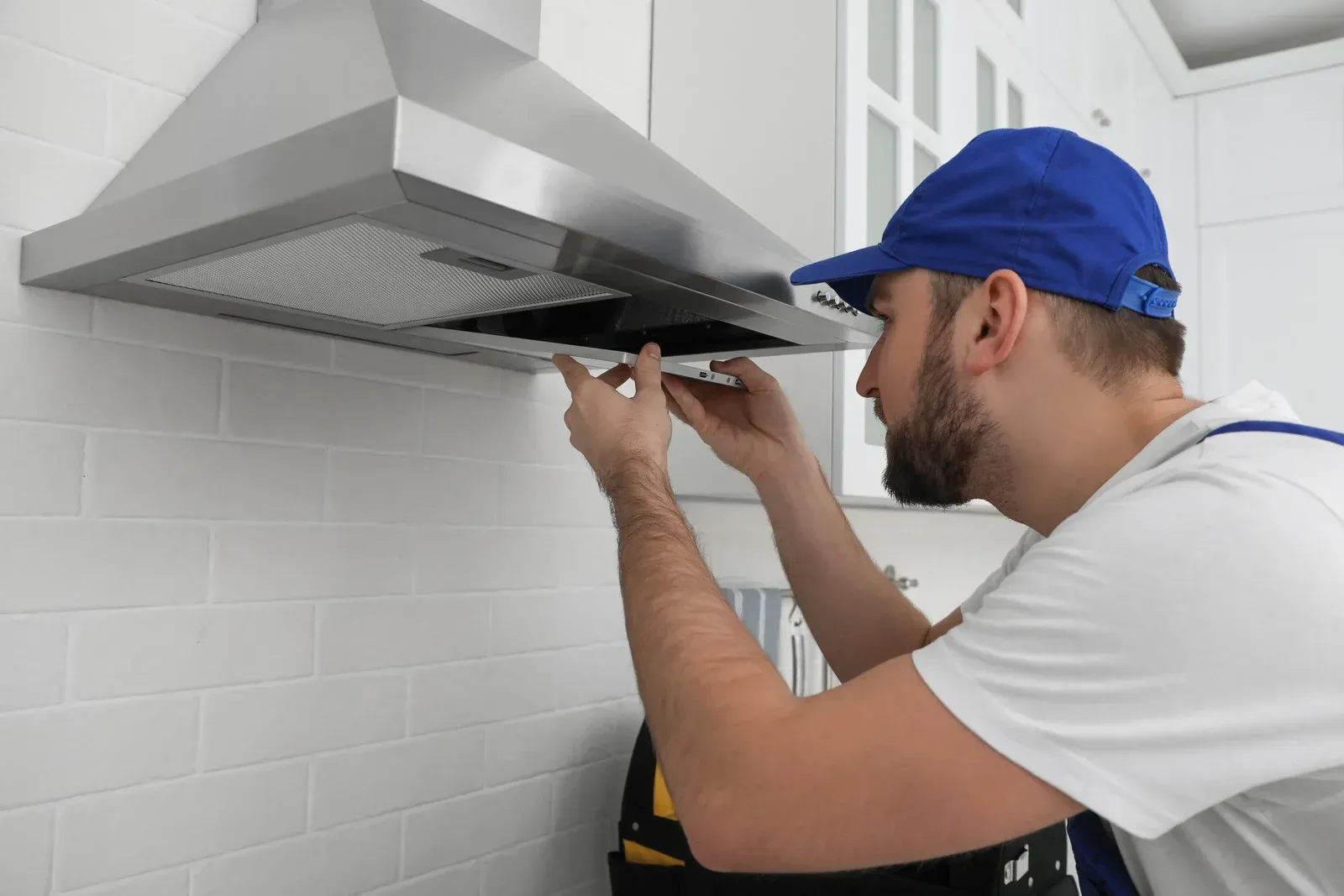 Man in blue cap installing a range hood in a white kitchen.