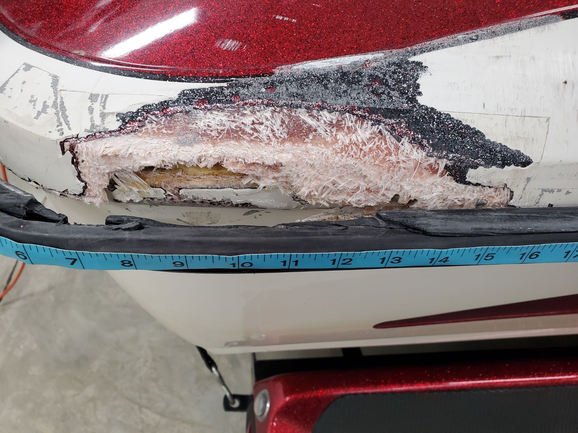 A close-up of a damaged fiberglass boat hull showing exposed wood and gelcoat layers above a black rub rail.