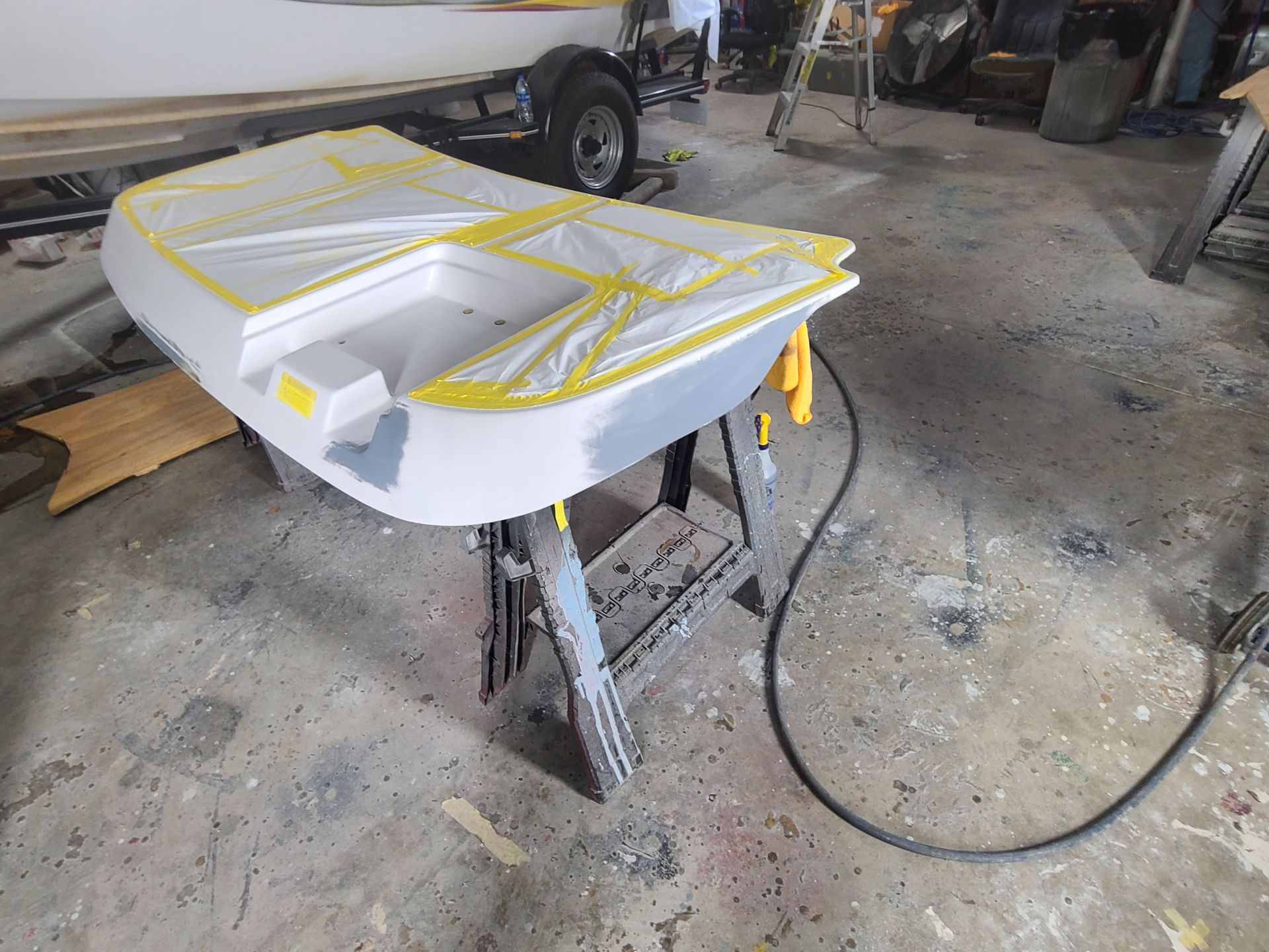 A boat part masked with yellow tape and partially sanded, resting on sawhorses in a workshop.