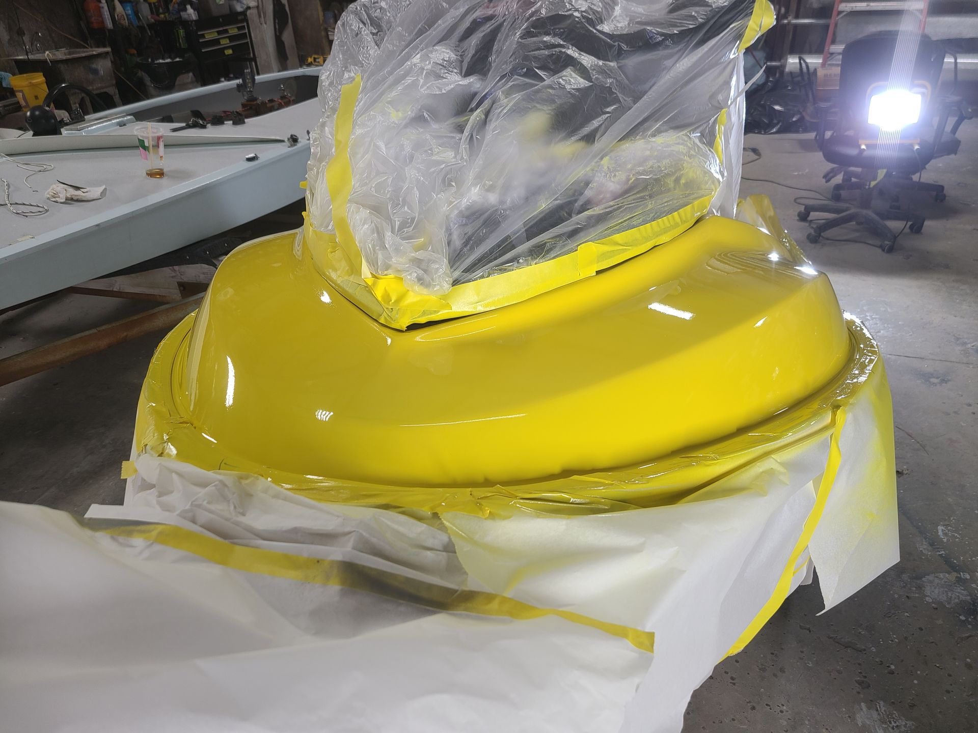 A bright yellow, freshly painted boat component partially covered in plastic and masking tape in a workshop.