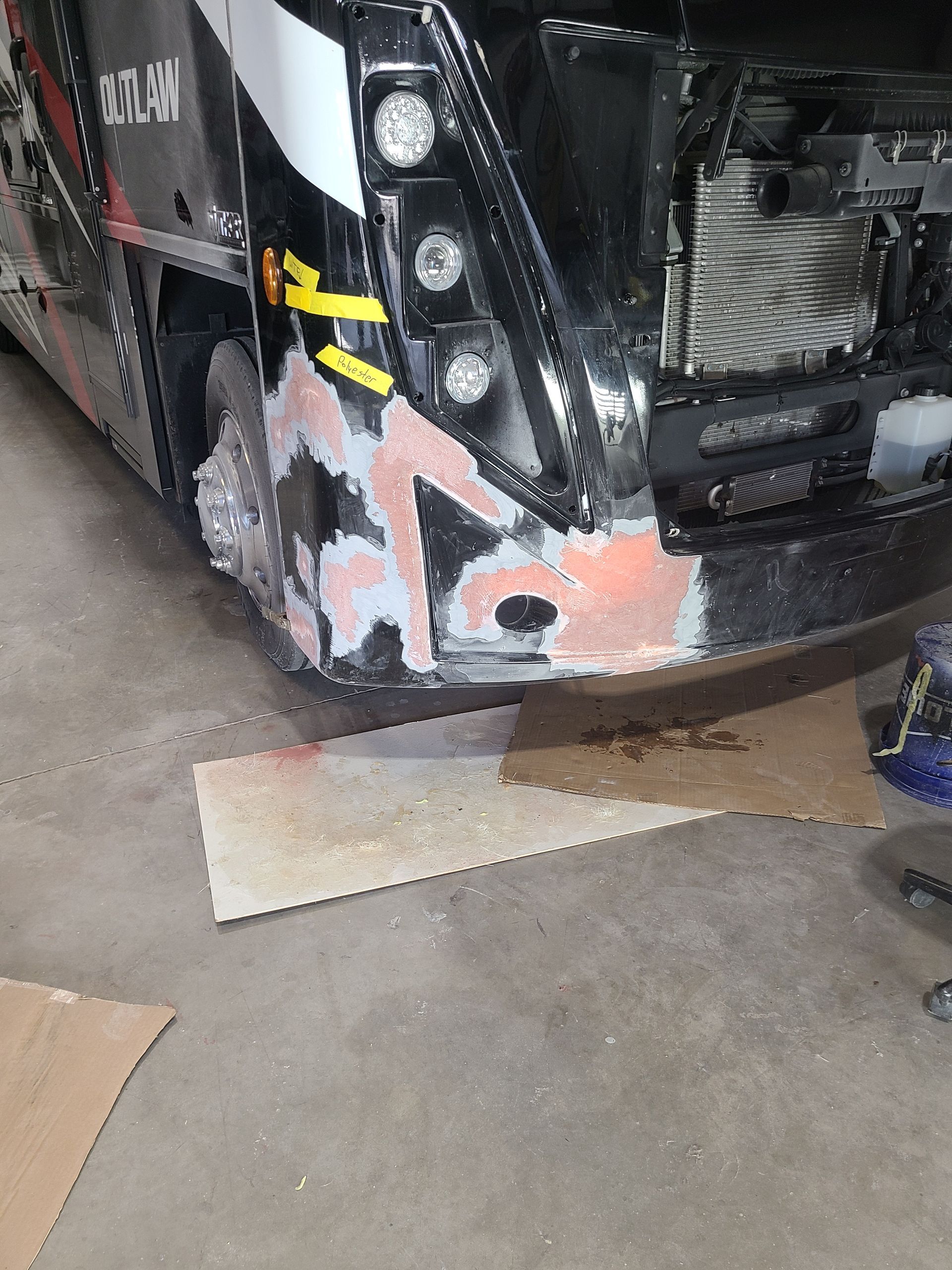 The front corner of a black bus undergoing auto body repair, showing sanded filler on the bumper in a garage setting.