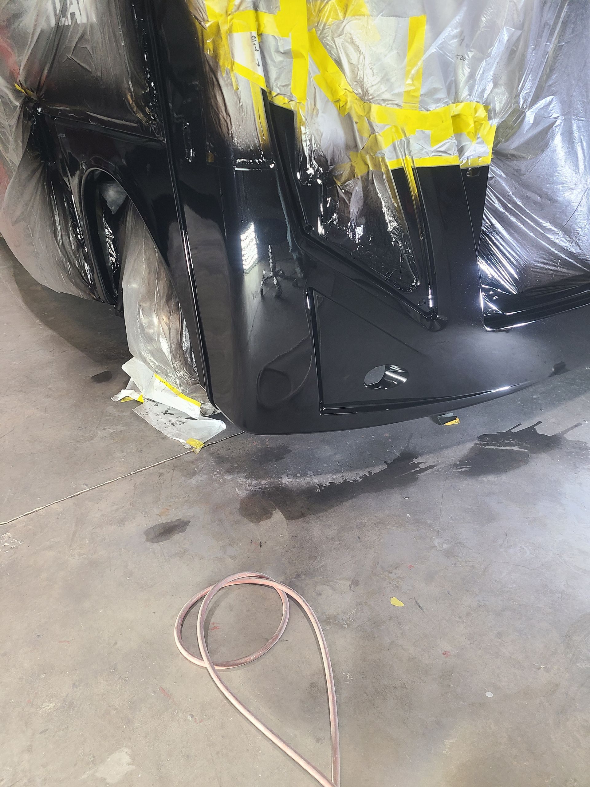A freshly painted glossy black bumper assembly sits on a concrete floor, partially covered by masking tape and plastic.