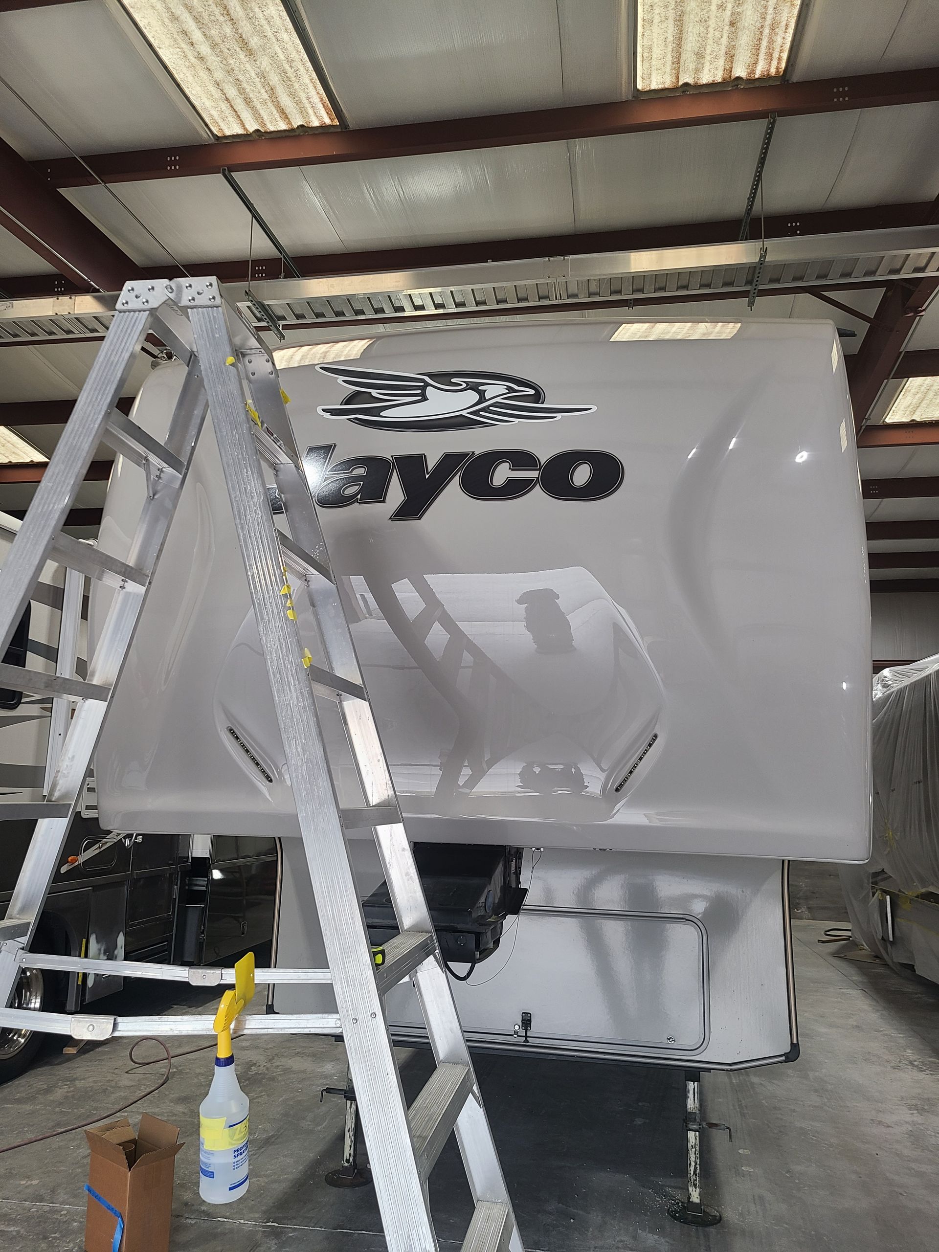 A silver ladder stands in front of the white, glossy front cap of a Jayco recreational vehicle inside a workshop.