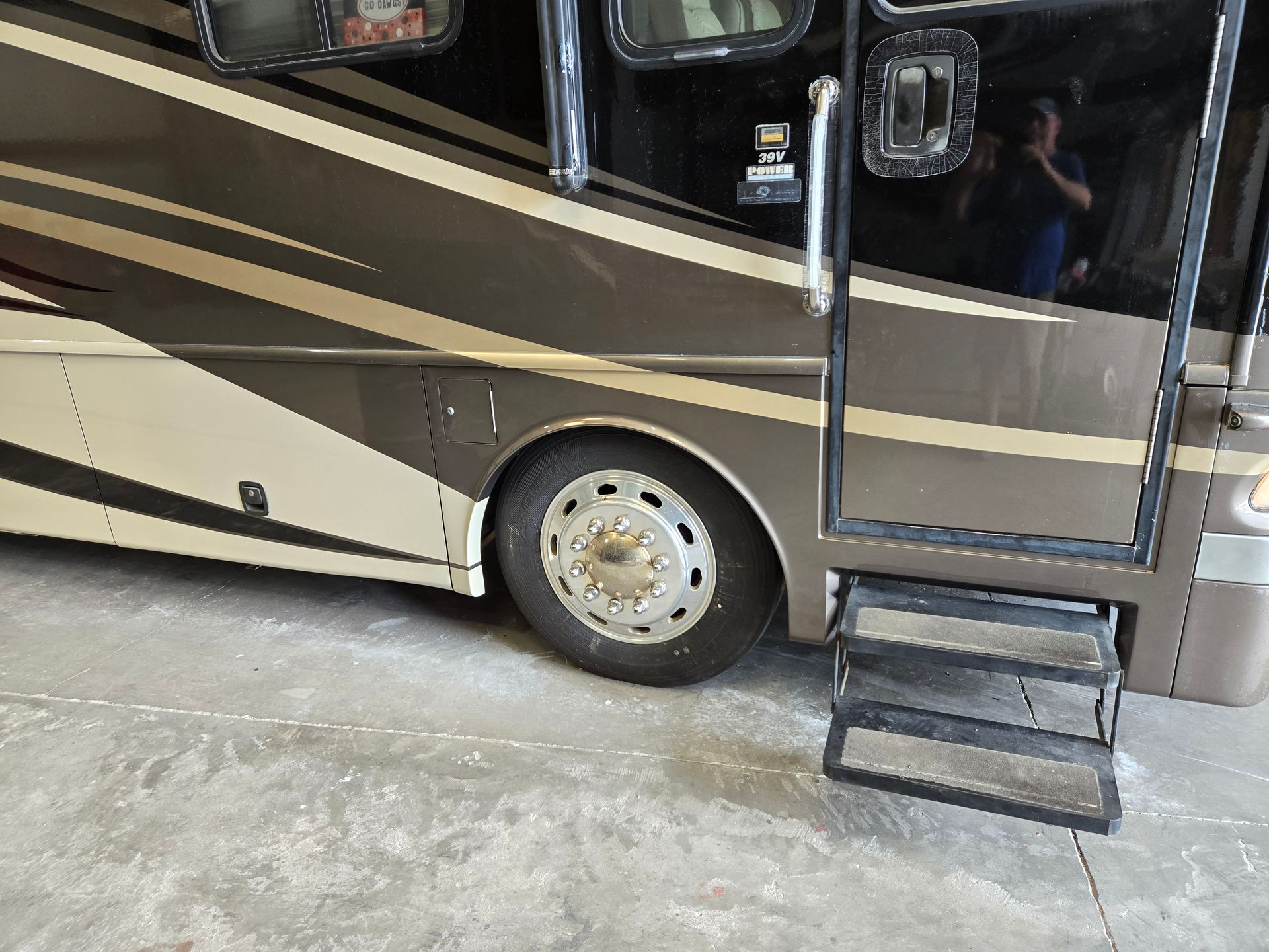 A parked RV with brown and tan siding, a front wheel, and extended metal stairs leading to the open passenger door.