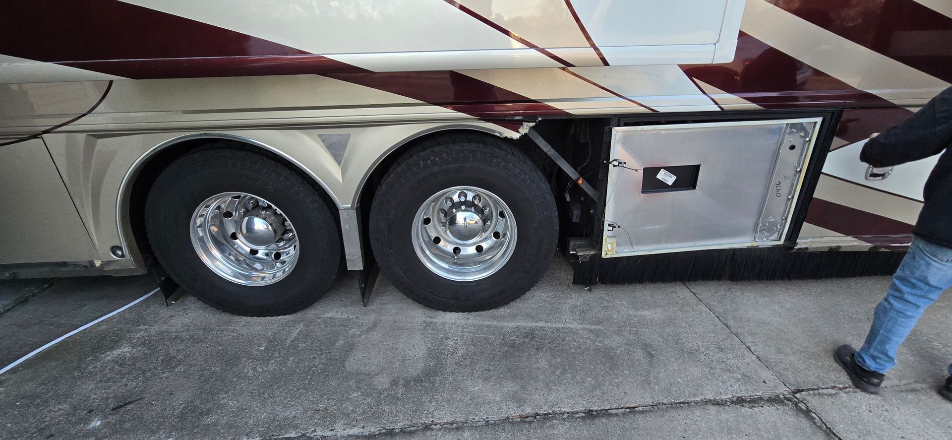 A side view of the rear wheels of a maroon and tan recreational vehicle with an open storage compartment door.