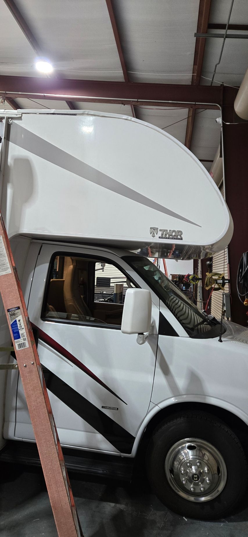 A side view of a white Thor Motor Coach RV parked inside a warehouse with a wooden ladder in the foreground.