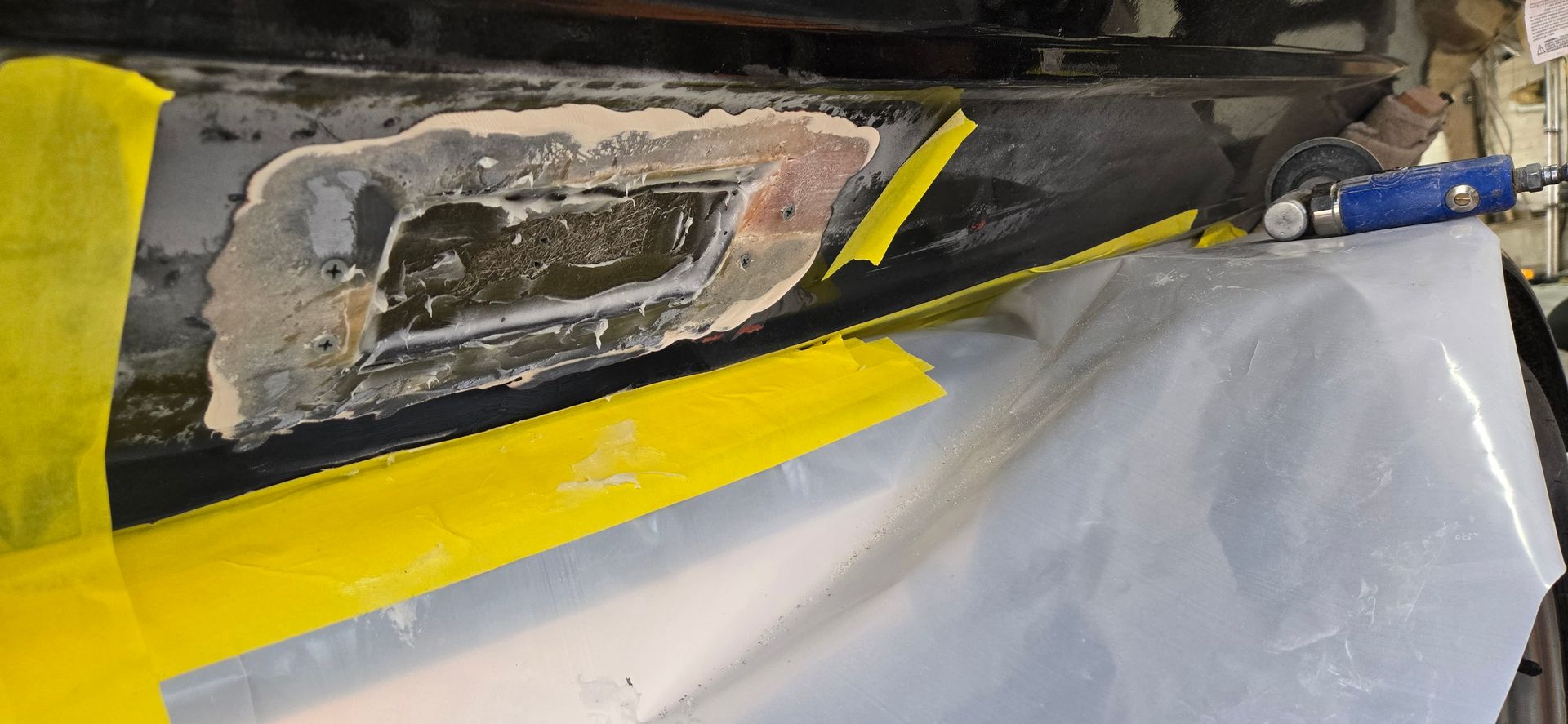 A damaged car rocker panel being prepared for repair, masked with yellow tape and covered by protective plastic.
