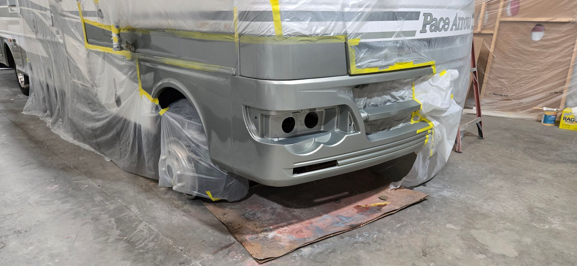 A Pace Arrow motorhome in a paint booth, with the front end freshly painted metallic gray and covered in masking plastic.