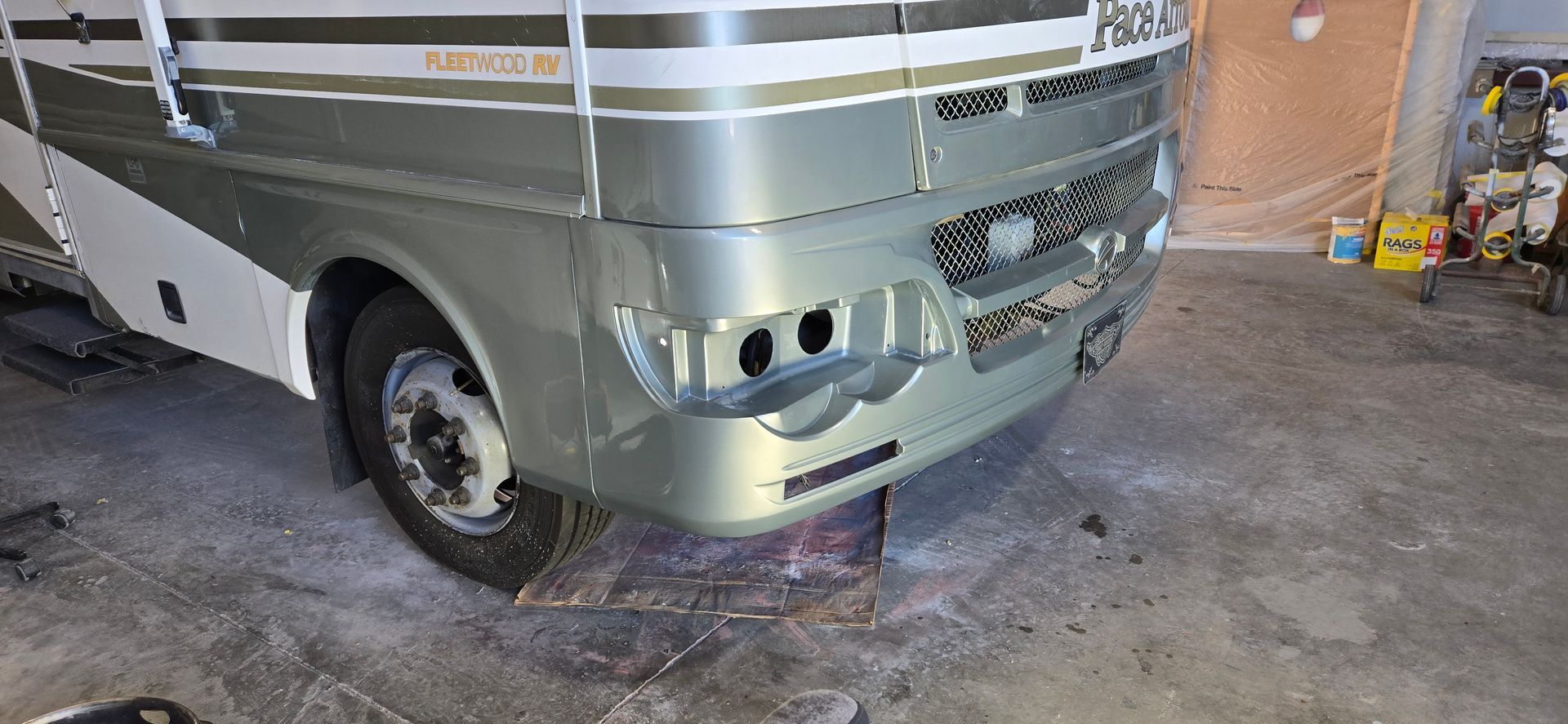 A green and white Fleetwood Pace Arrow RV in a repair shop with its front headlights and grille disassembled.