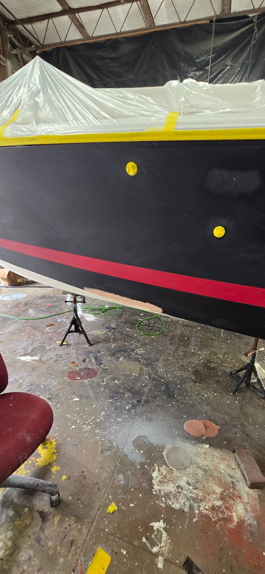 A boat hull painted black with a red stripe and yellow markers, suspended on jack stands in a workshop.