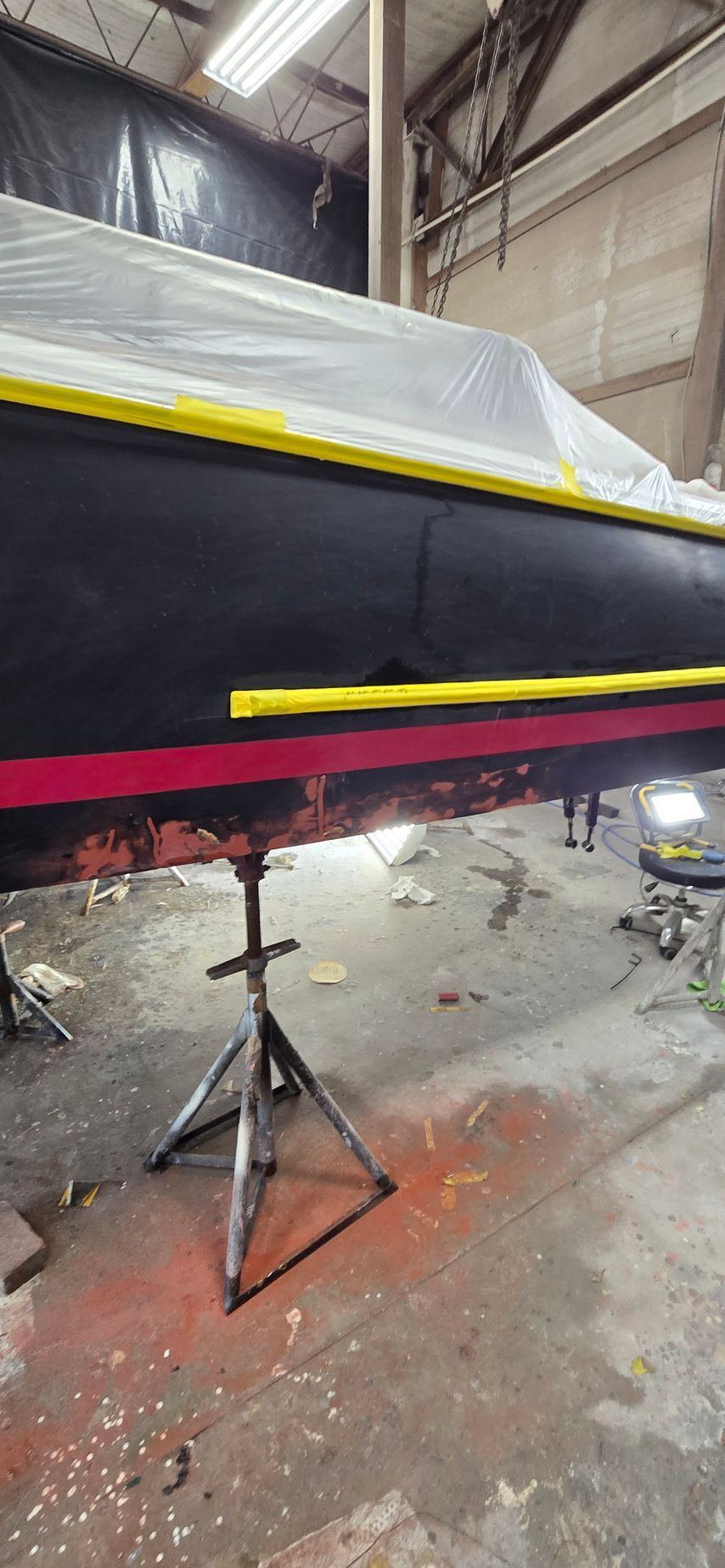 A boat hull on a jack stand in a workshop, featuring masked black and red sections with yellow tape.