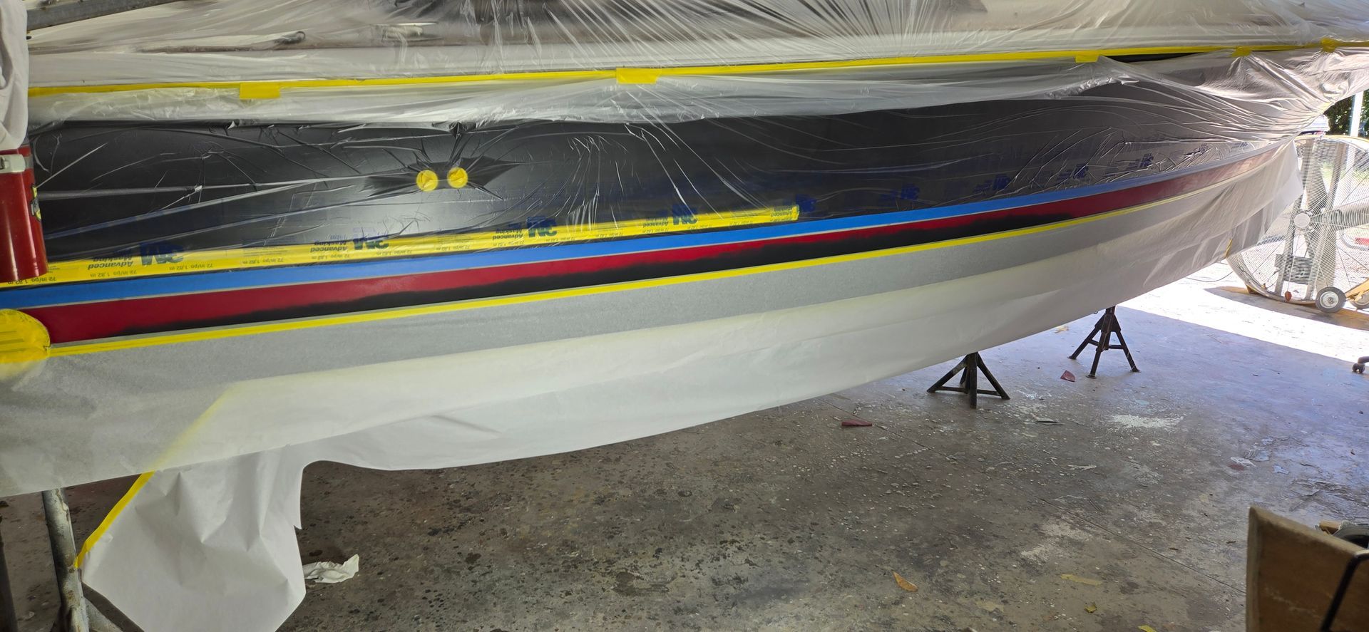 A boat hull in a workshop, masked with tape and plastic for painting, featuring black, blue, red, and grey sections.