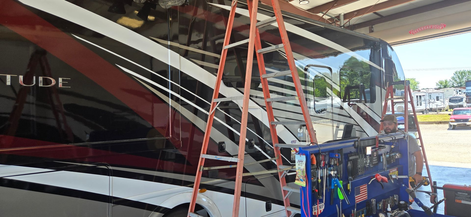 A technician works on the exterior of a large, red and white recreational vehicle inside a service bay.