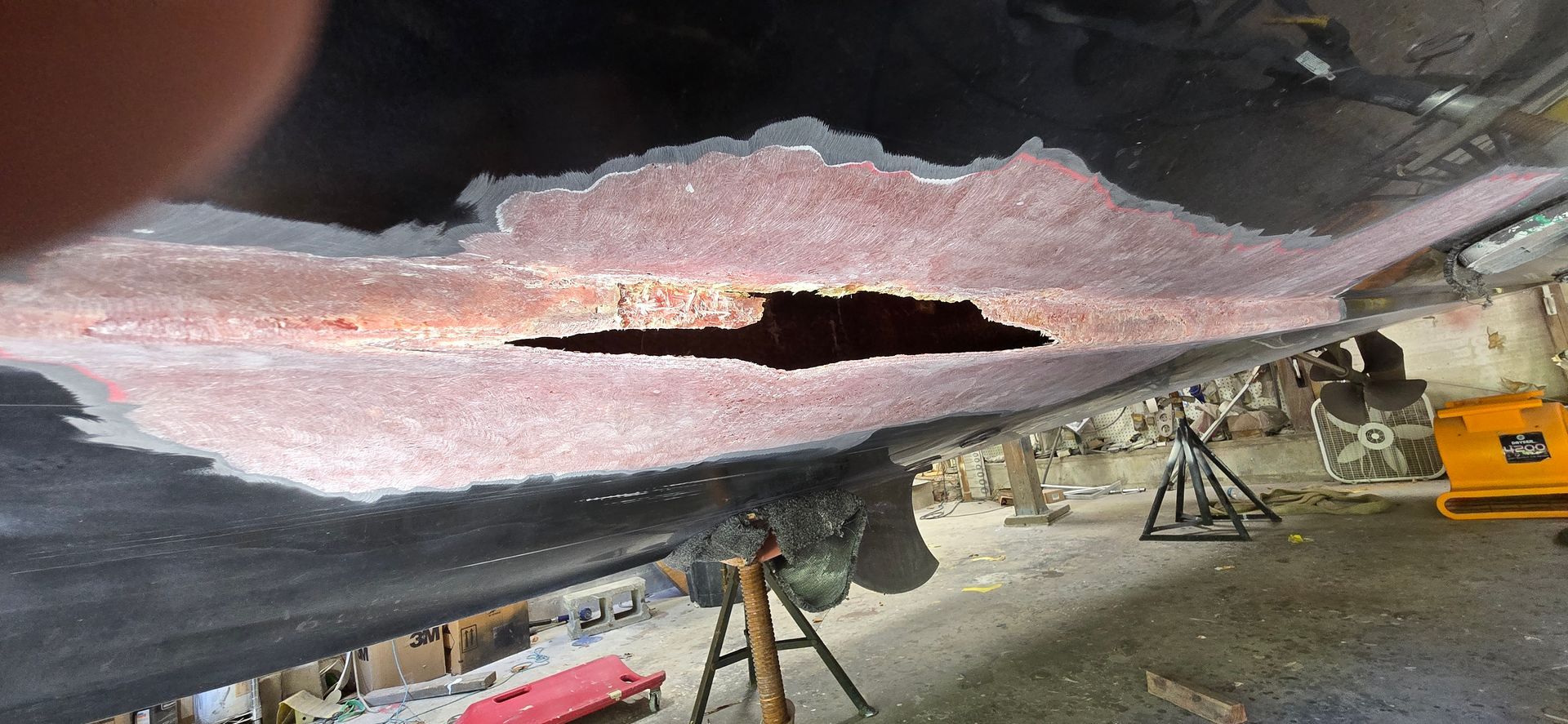 A large, jagged hole in the underside of a boat hull, exposing the inner fiberglass structure within a workshop.