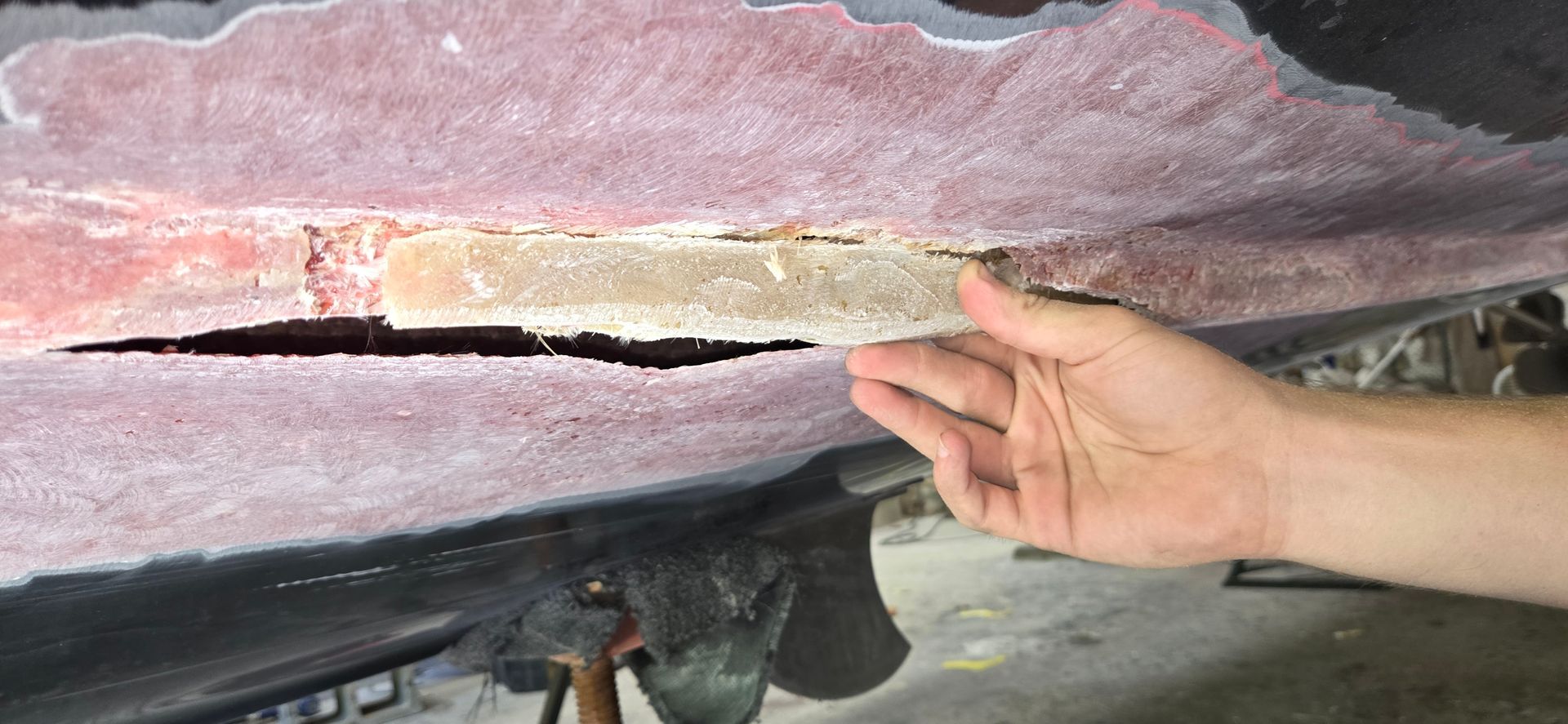 A hand holds a piece of foam core exposed by a large crack in the fiberglass hull of a boat in a workshop.