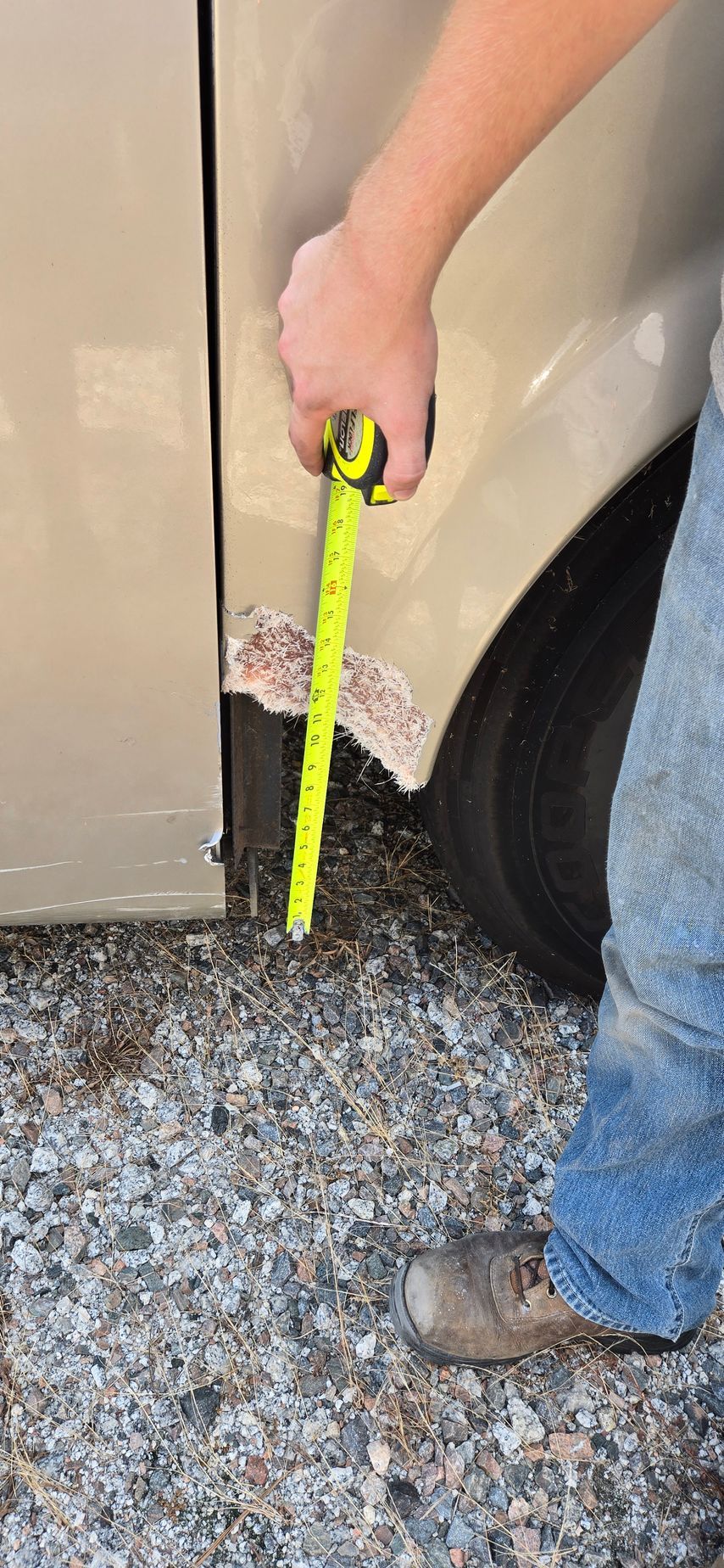 A person measures a patch of rust on the lower wheel arch of a tan vehicle using a yellow tape measure.