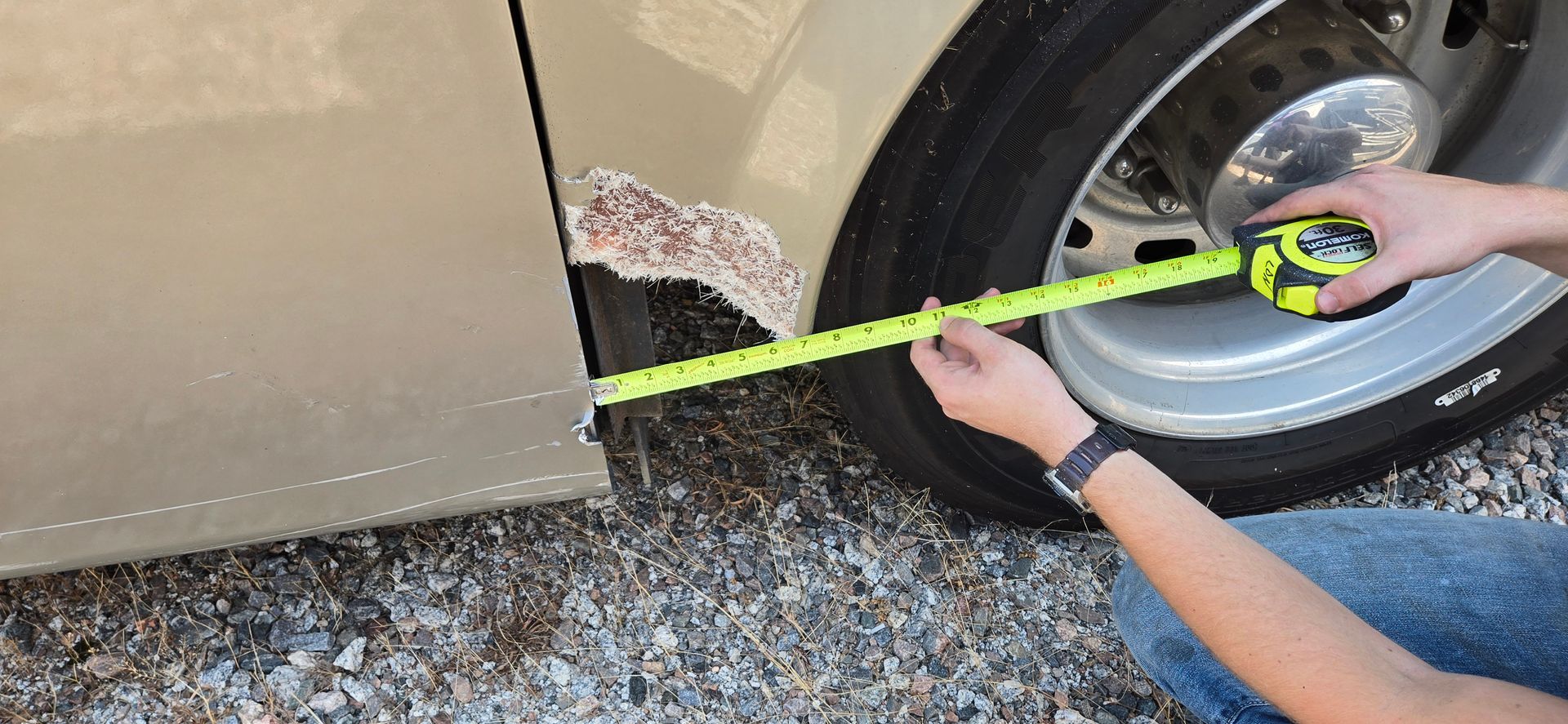 A person holds a yellow tape measure against a beige vehicle's rusted wheel well to determine the size of the area.