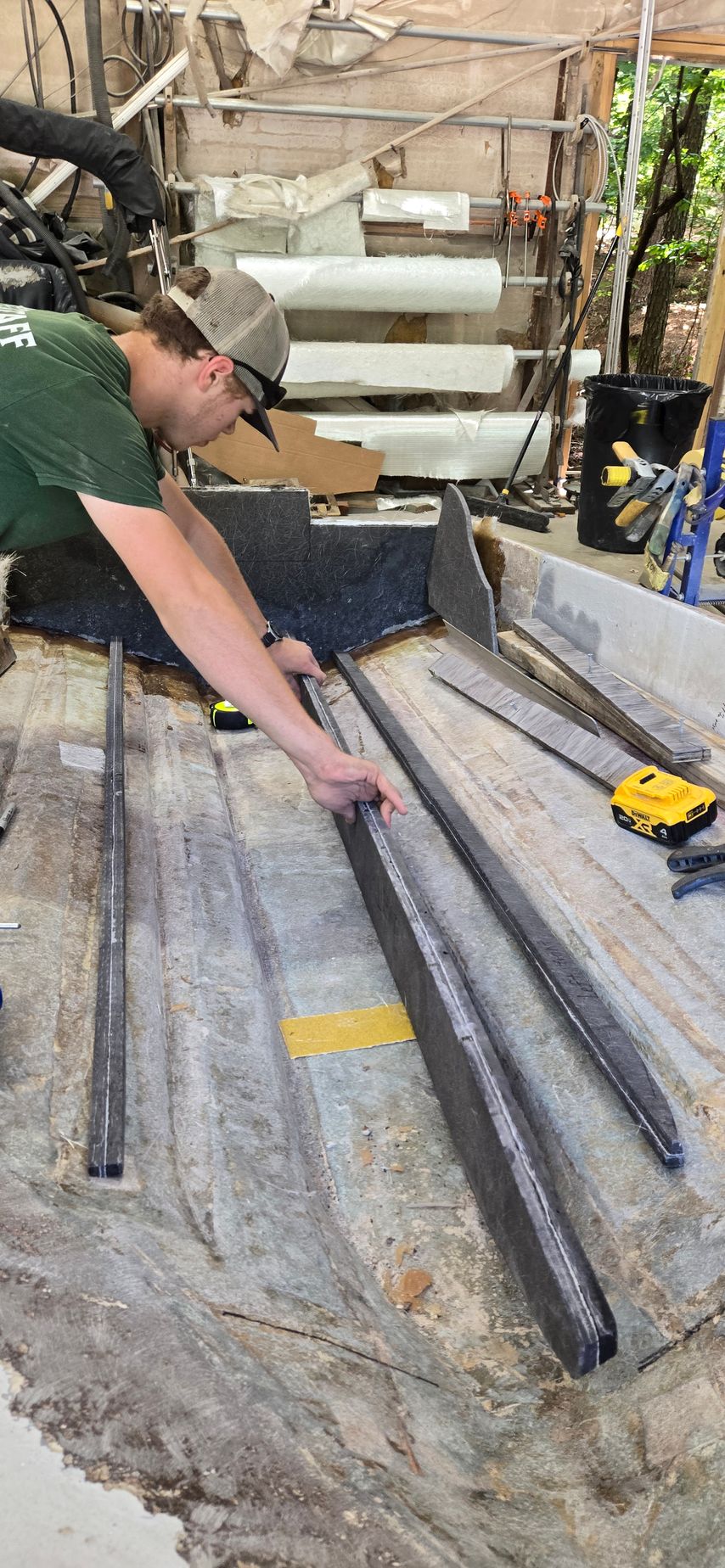 A person wearing a hat and green shirt measures metal framing strips on a workspace floor.