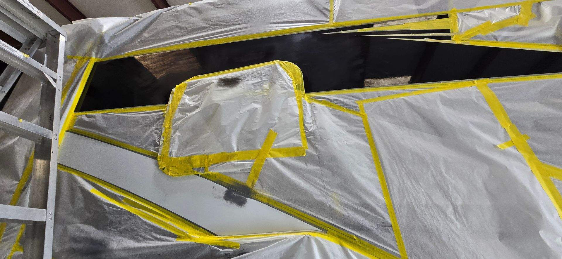 A car part being prepared for painting, covered in protective white plastic sheeting held down by yellow masking tape.