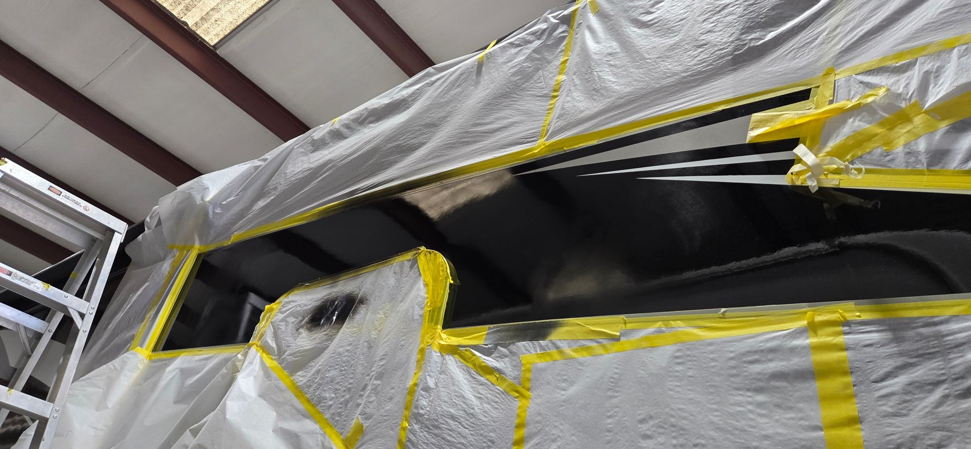 Vehicle parts masked with plastic sheeting and yellow tape in an auto body shop.