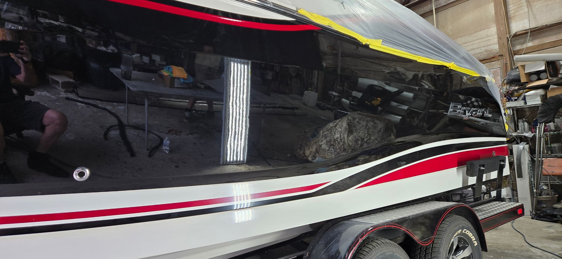 A person polishes the reflective black hull of a boat on a trailer inside a workshop, with a bright light reflecting on it.