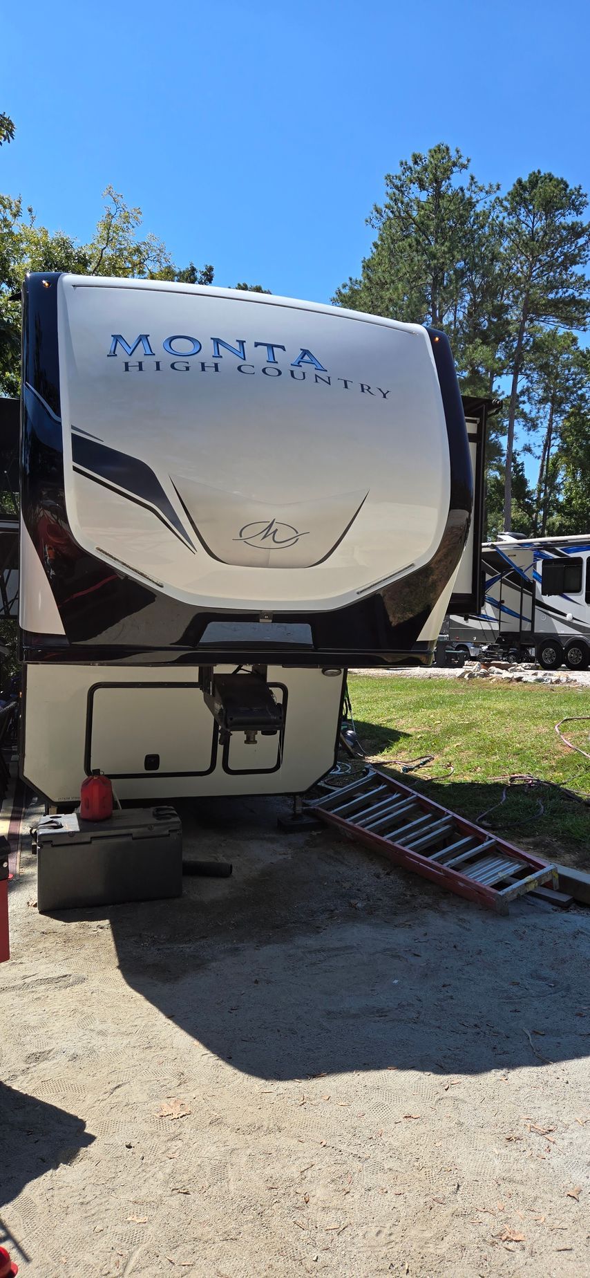 A white and black Montana High Country fifth-wheel travel trailer parked on a gravel lot under a clear blue sky.