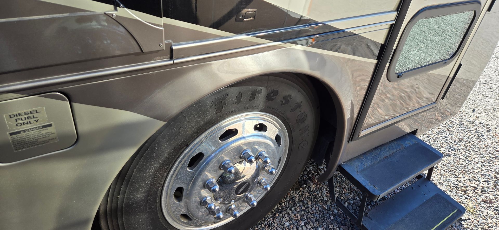 A close-up view of a motorhome’s wheel, polished chrome rim, fuel cap, and a set of retracted steps on a gravel surface.