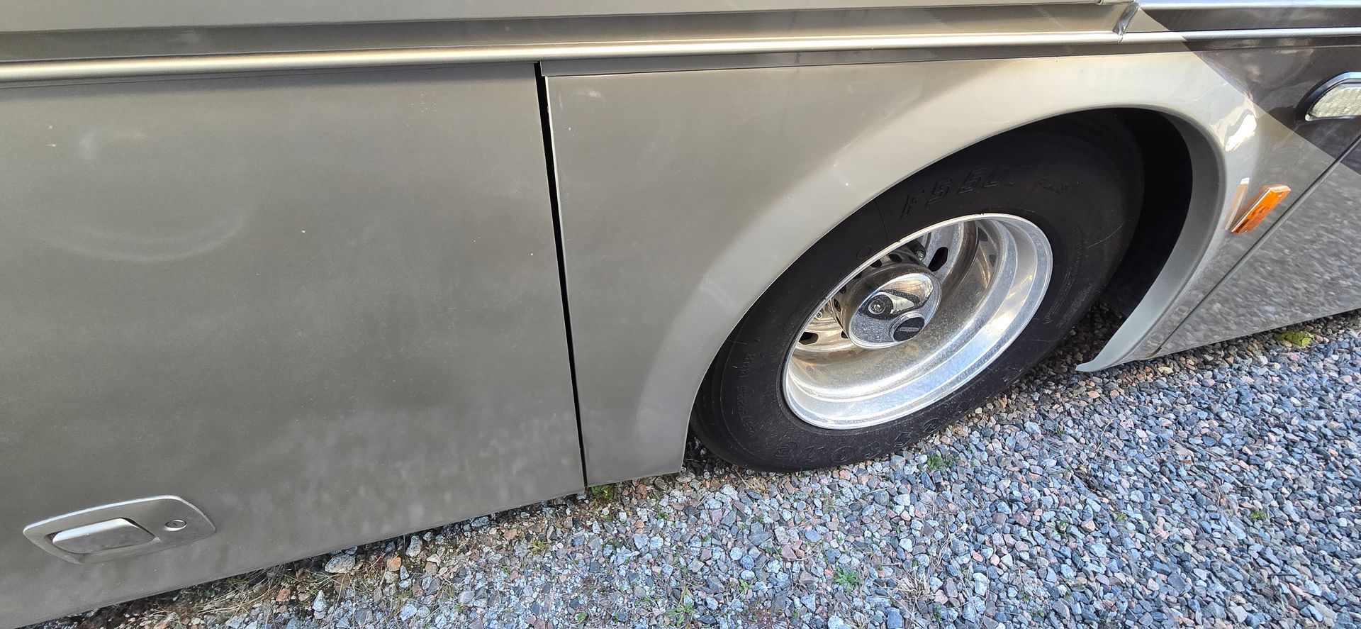 A close-up view of the side of a grey motorhome, showing a wheel and tire parked on gravel.