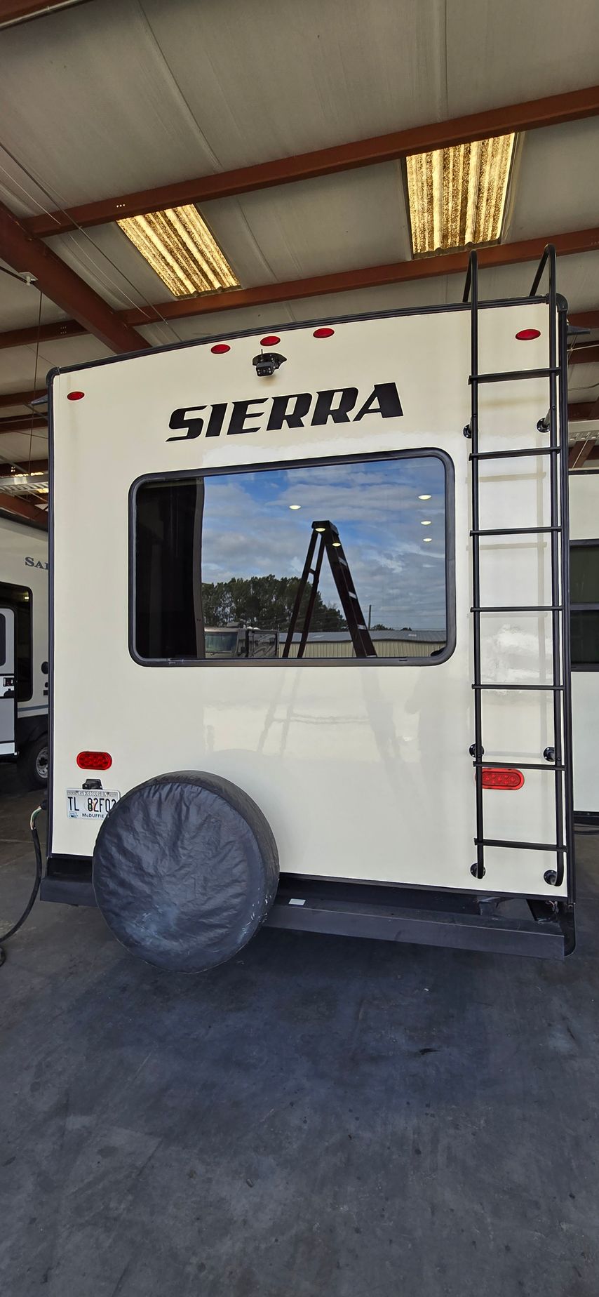 The back of a cream-colored Sierra RV inside a warehouse, featuring a window, ladder, and a covered spare tire.