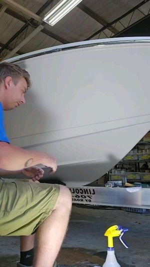 A person wearing green shorts works on the side of a large boat in a workshop with a spray bottle in the foreground.