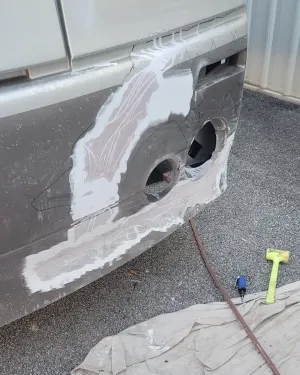 A car bumper in the process of repair, featuring body filler applied over surface damage near two circular openings.