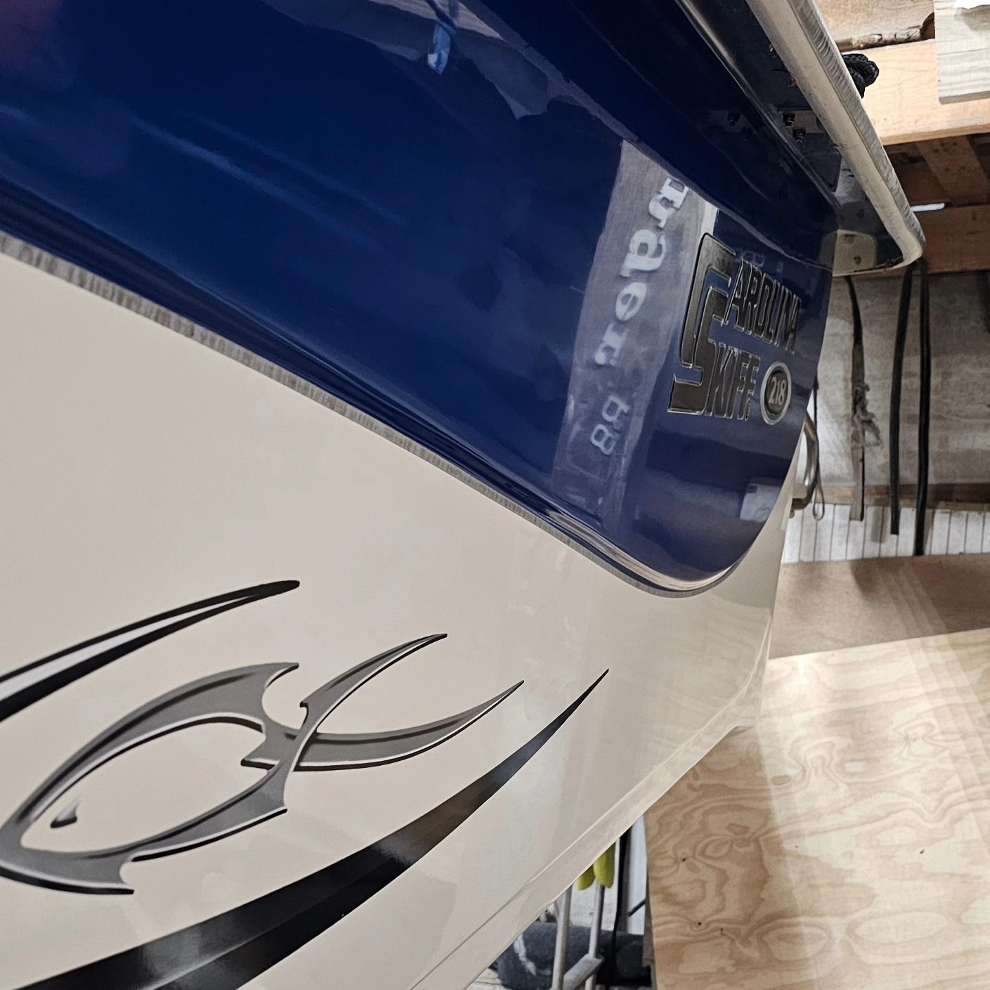 Close-up of a blue and off-white boat hull featuring a stylized fish decal, seen at an angle in a workshop setting.