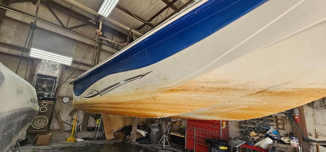 A boat hull with a blue stripe on top and a stained, tan-colored lower portion, suspended inside a repair workshop.