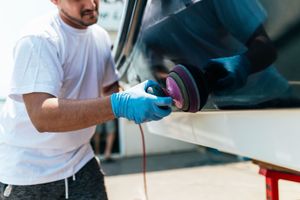 A person wearing blue gloves uses a rotary polisher to buff the dark, reflective surface of a boat.