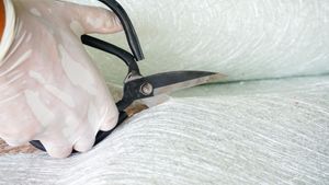 A gloved hand uses heavy-duty shears to cut a sheet of white fiberglass matting.