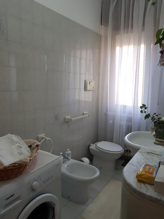 Bagno con WC, bidet, lavatrice e finestra con tende trasparenti. Pareti piastrellate bianche e lavabo in marmo.